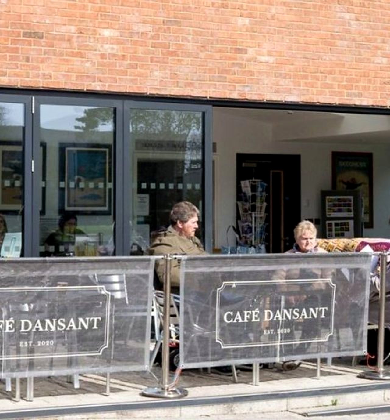The front of Café Dansant: brick pavilion with tall glass doors, customers at tables behind dark screens printed with the café’s name