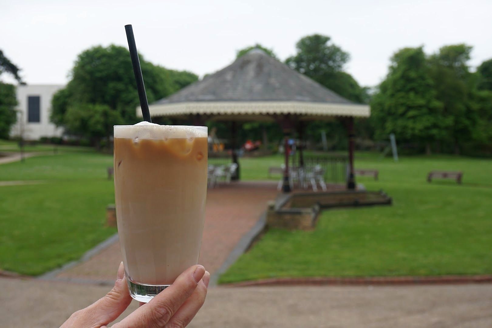 An iced coffee held up in front of the lawns and the bandstand in Tower Gardens