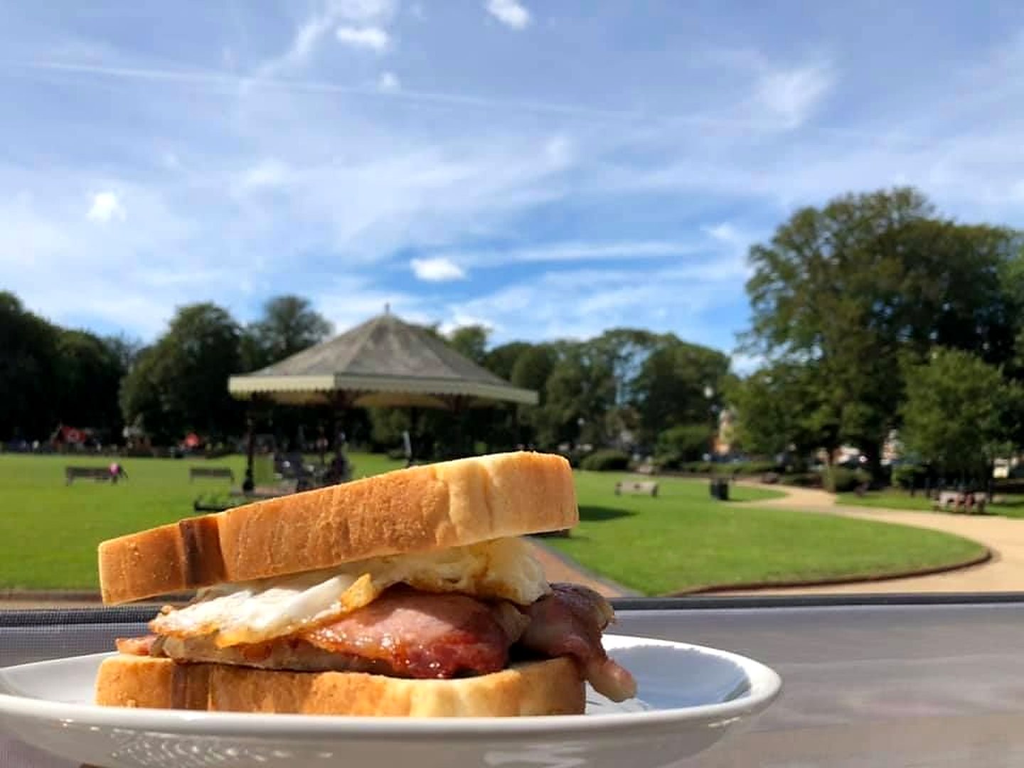 A bacon and egg sandwich on white bread, on a plate outside with the bandstand behind